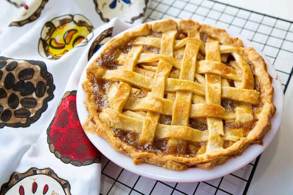 A lattice-topped pie in a white dish is cooling on a wire rack, next to a cloth with colorful pie illustrations.
