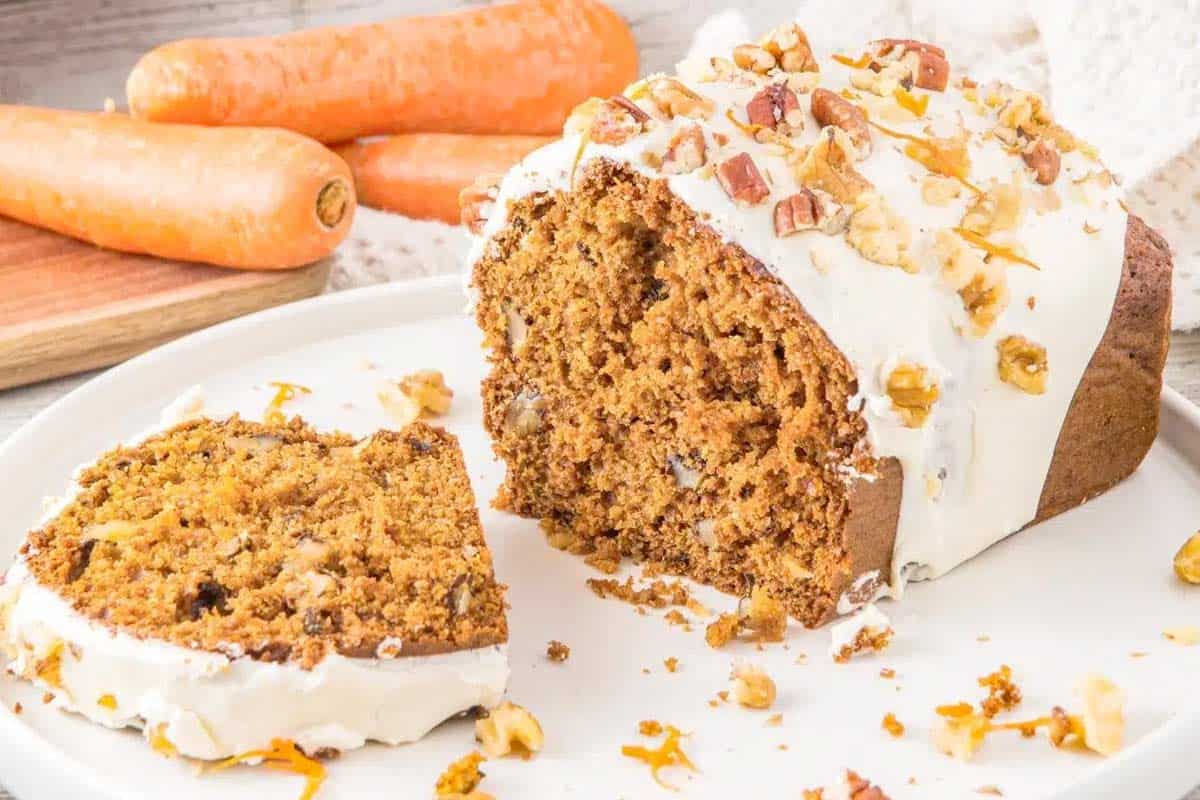 A loaf of carrot cake with white frosting and chopped nuts on top, partially sliced, sits on a white plate with whole carrots in the background.