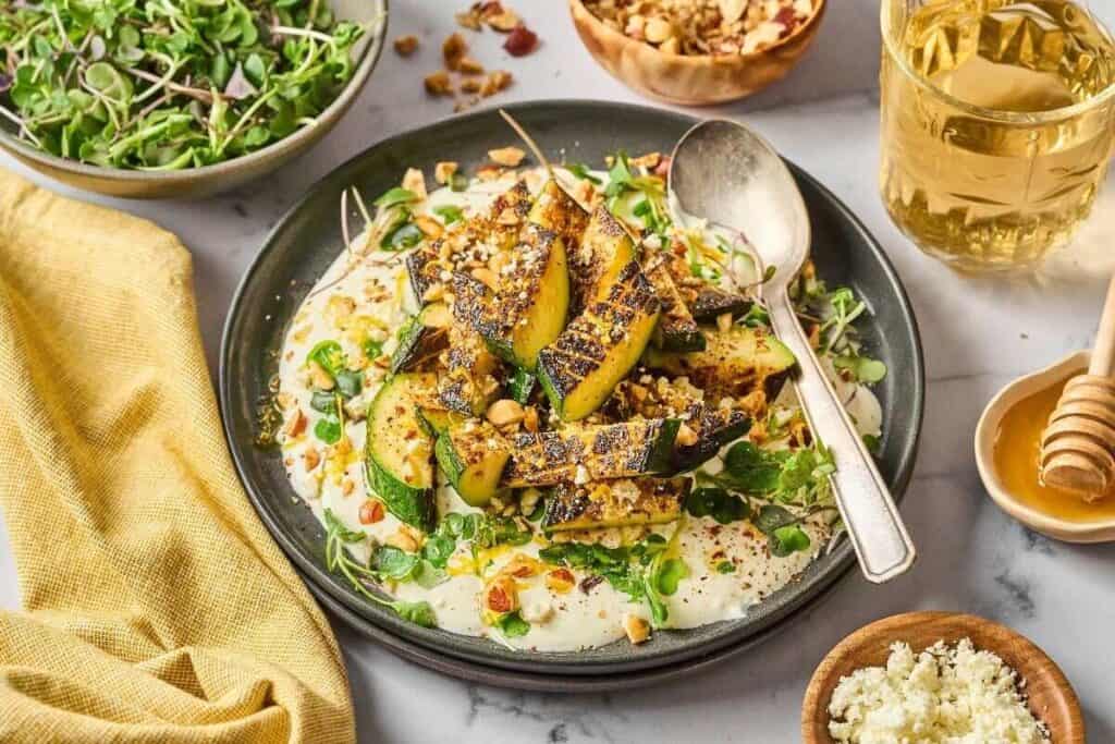 Grilled zucchini slices sit on a bed of creamy sauce, topped with chopped nuts and greens, with drinks and various toppings in bowls nearby.