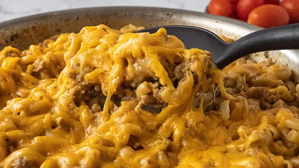 Close-up of a skillet filled with cheesy ground meat and pasta casserole, with a black spoon scooping out a portion.