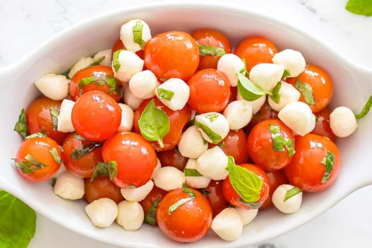 A white dish filled with cherry tomatoes, small mozzarella balls, and fresh basil leaves.