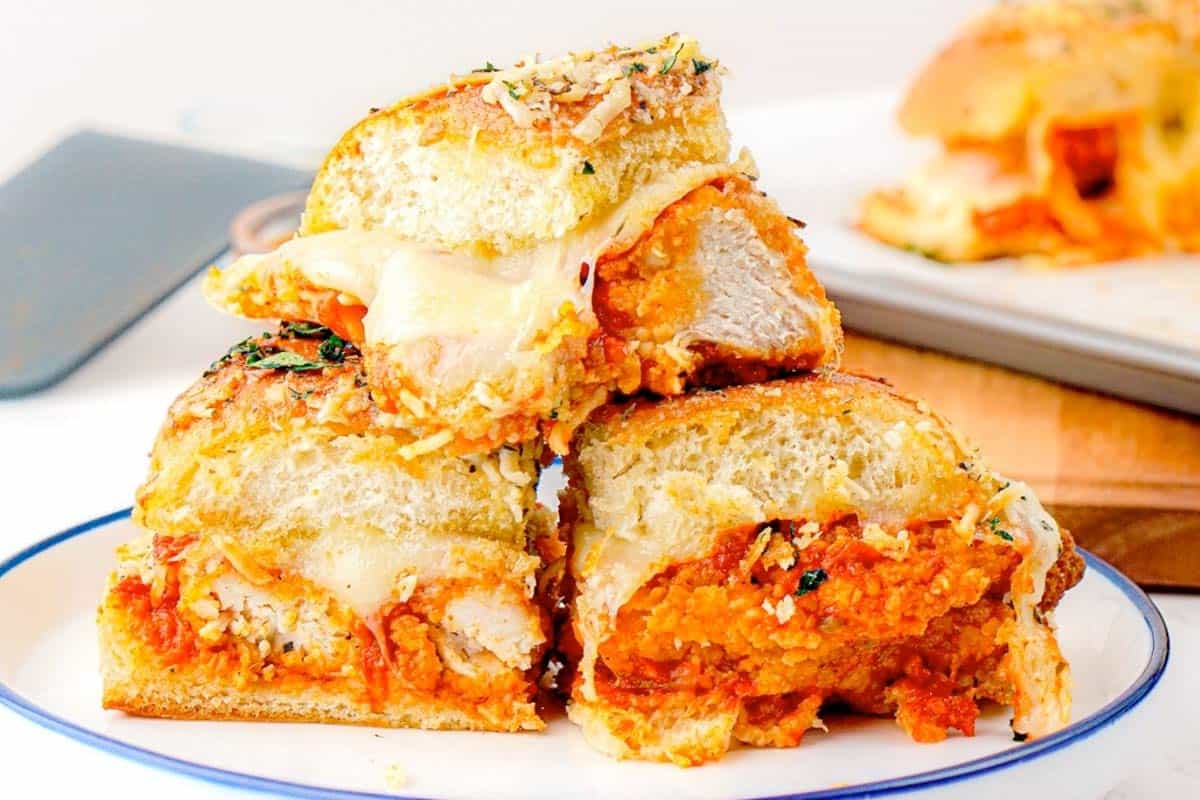A plate with three stacked Chicken Parmesan Sliders, featuring breaded meat, tomato sauce, and melted cheese, with a baking tray and phone in the background.