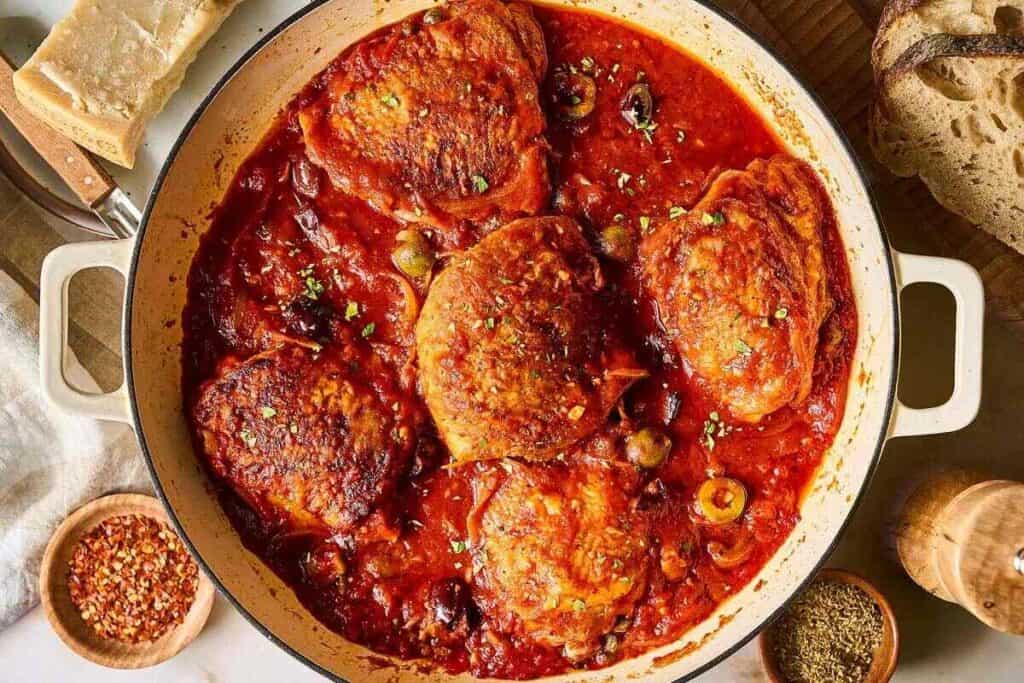 A pot of braised chicken thighs in tomato sauce with olives and herbs, surrounded by bread, grated cheese, and small bowls of spices.