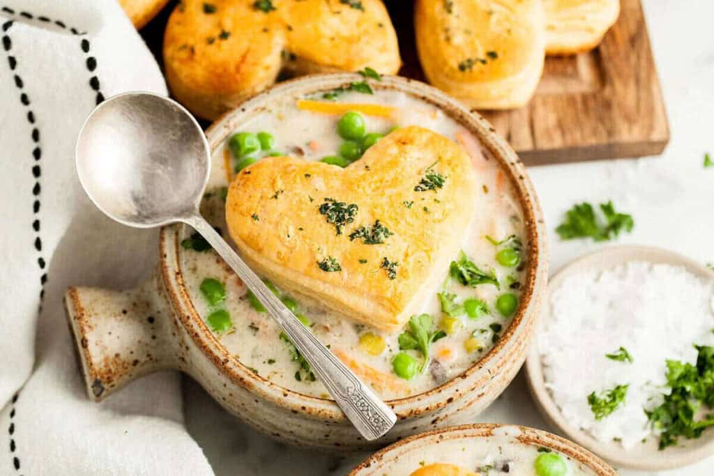 A bowl of creamy soup with peas, topped with a heart-shaped biscuit and garnished with herbs, next to a spoon and a tray of more biscuits.
