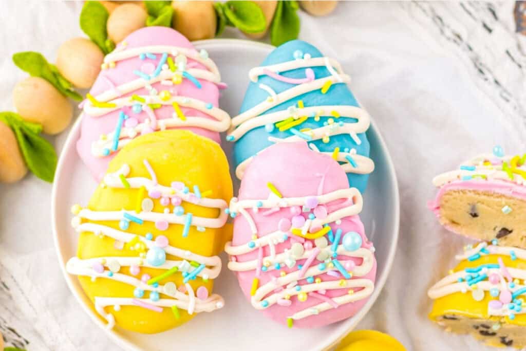 A plate of pastel-colored, egg-shaped treats with icing drizzle and sprinkles; one treat is cut open showing a cookie dough filling.