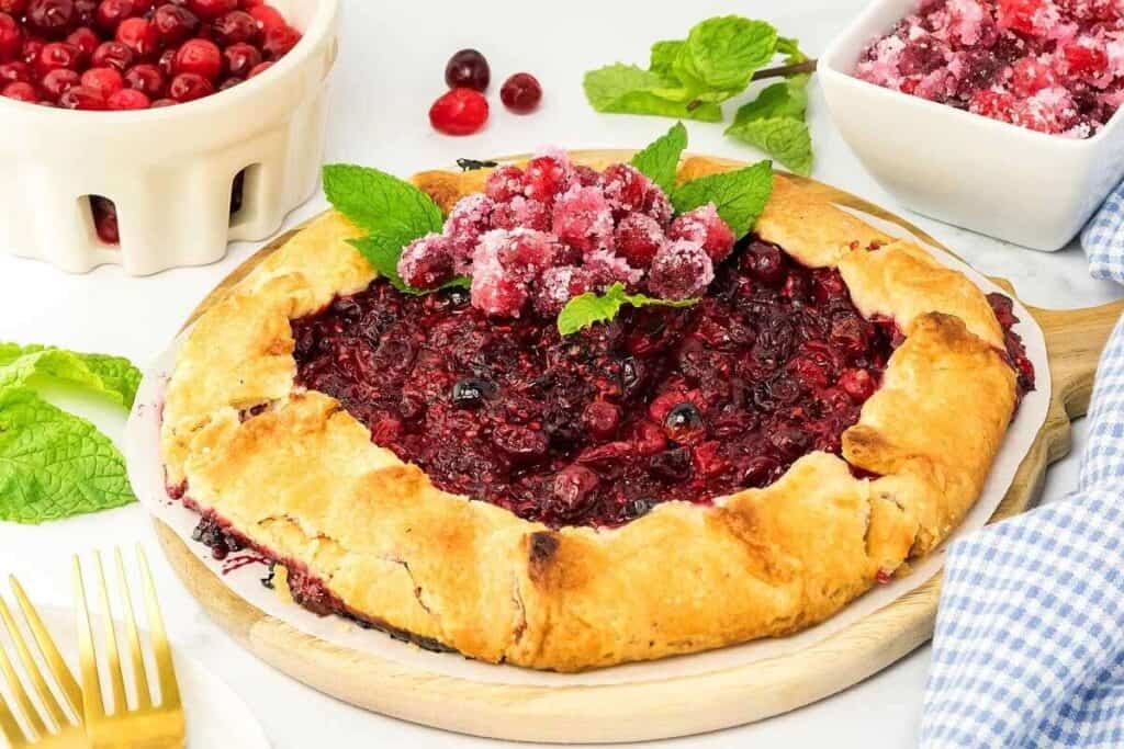 A rustic berry galette with a golden crust, topped with sugared cranberries and mint leaves, sits on a wooden board next to bowls of cherries and utensils.