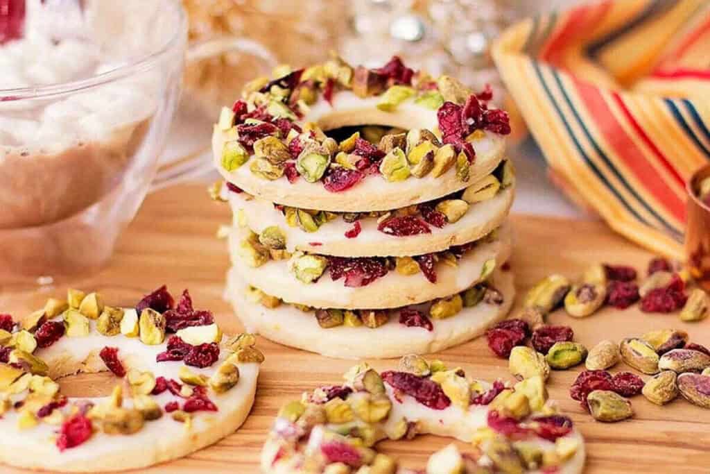 A stack of round cookies topped with chopped pistachios and dried cranberries is displayed on a wooden surface next to a striped napkin.