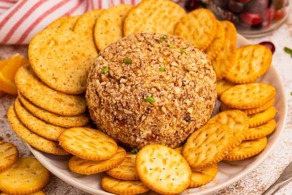 A round cheese ball coated in chopped nuts is surrounded by assorted crackers on a white plate.