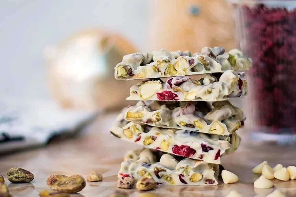A stack of white chocolate bark pieces with pistachios and dried cranberries, surrounded by loose nuts and chocolate chips.