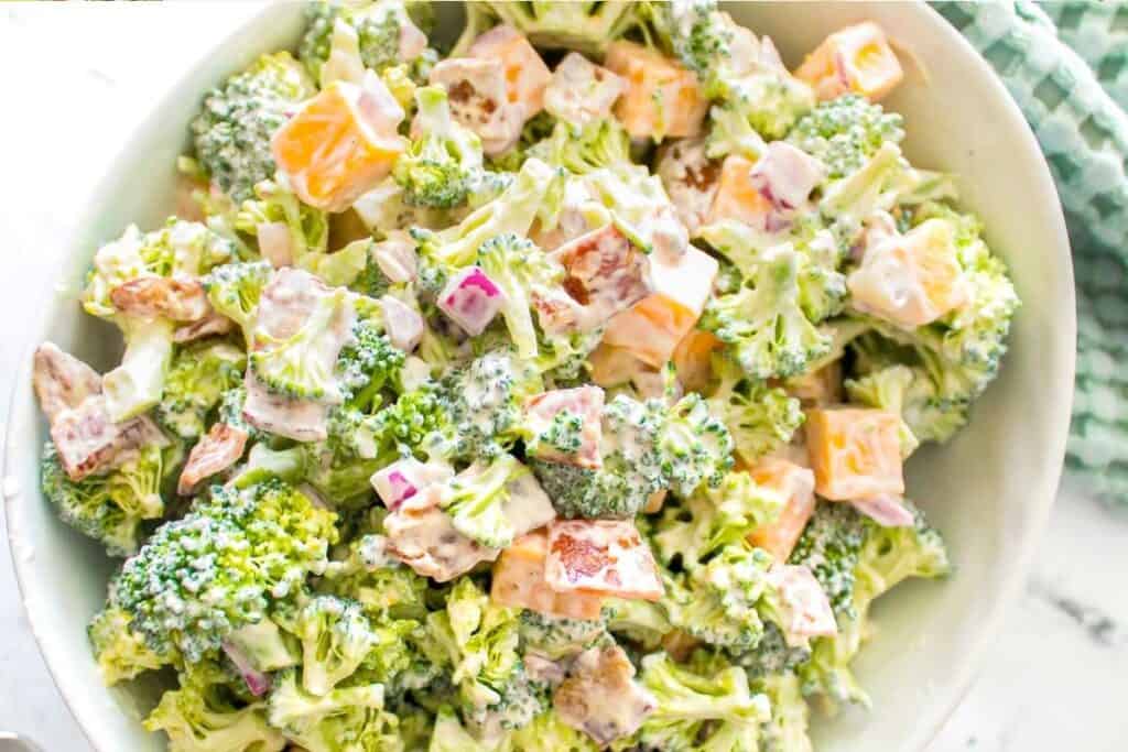 A bowl of broccoli salad with chopped broccoli, cheddar cheese cubes, red onion, bacon pieces, and creamy dressing.
