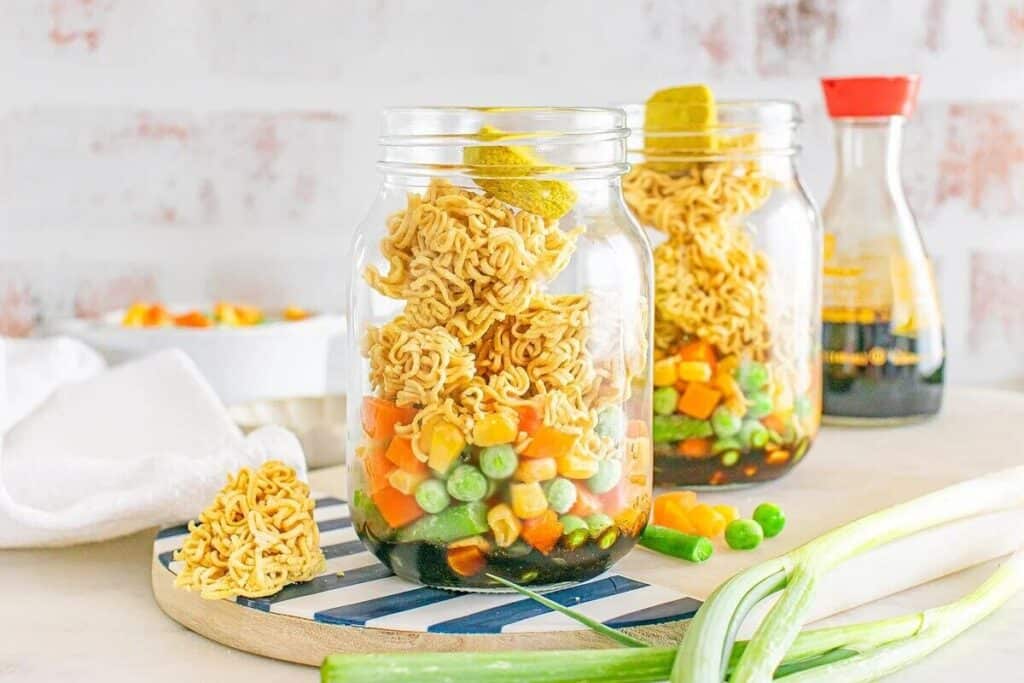 Two mason jars filled with uncooked instant noodles, mixed vegetables, and broth base, with scallions and a soy sauce bottle nearby on a kitchen counter.