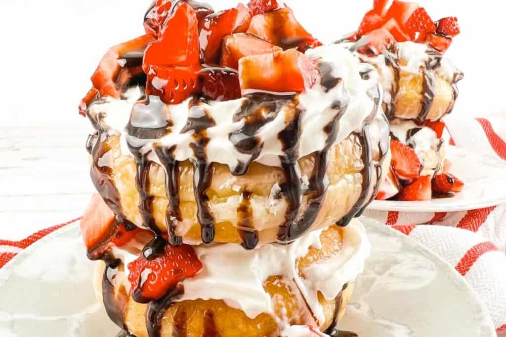 Two stacked donuts topped with whipped cream, sliced strawberries, and chocolate drizzle on a white plate.