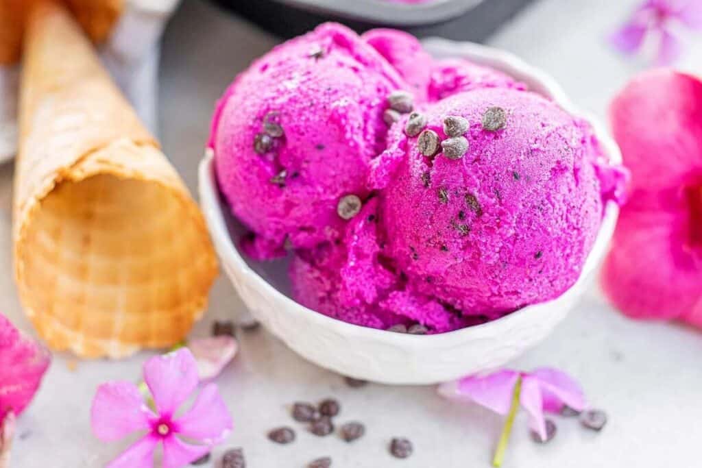 A white bowl filled with two scoops of bright pink ice cream topped with chocolate chips, next to an empty waffle cone and pink flowers.