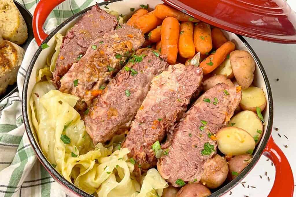 A pot filled with sliced corned beef, baby carrots, cabbage, and halved red potatoes, garnished with chopped herbs.