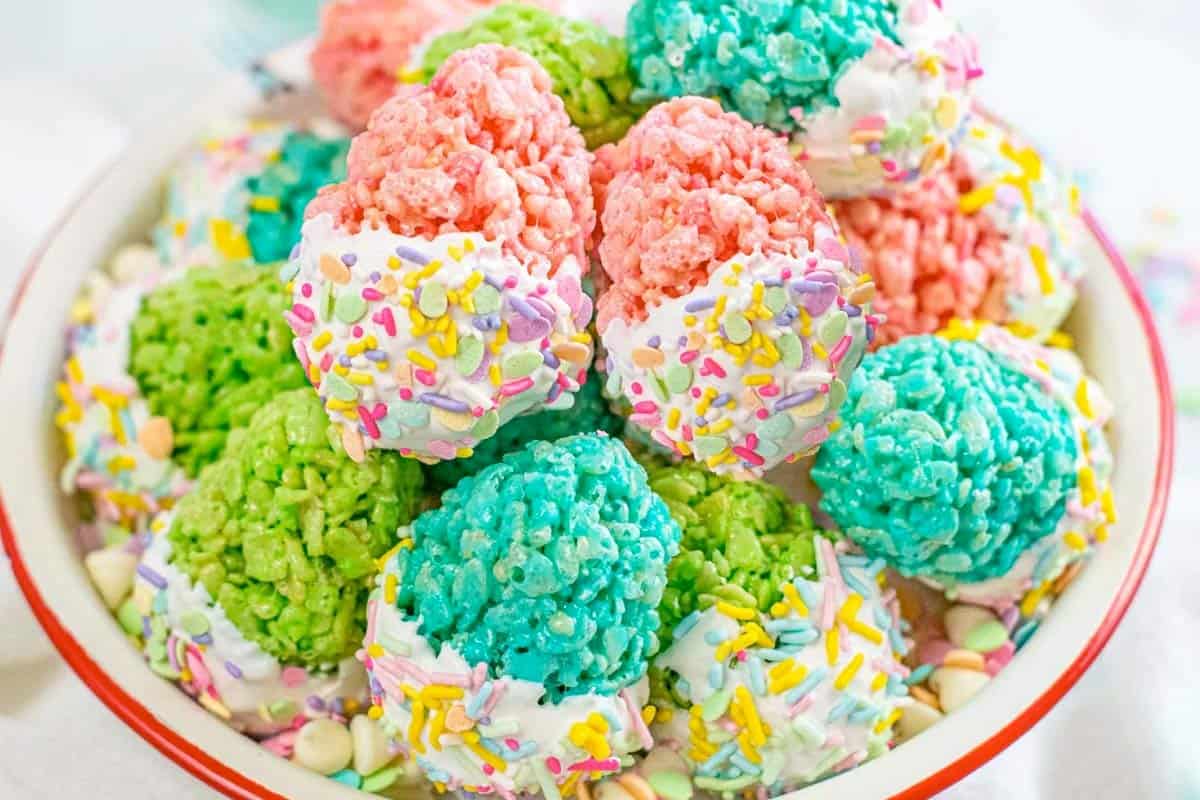 A bowl filled with colorful rice krispie treats in pink, green, and blue, each dipped in white chocolate and decorated with assorted sprinkles.