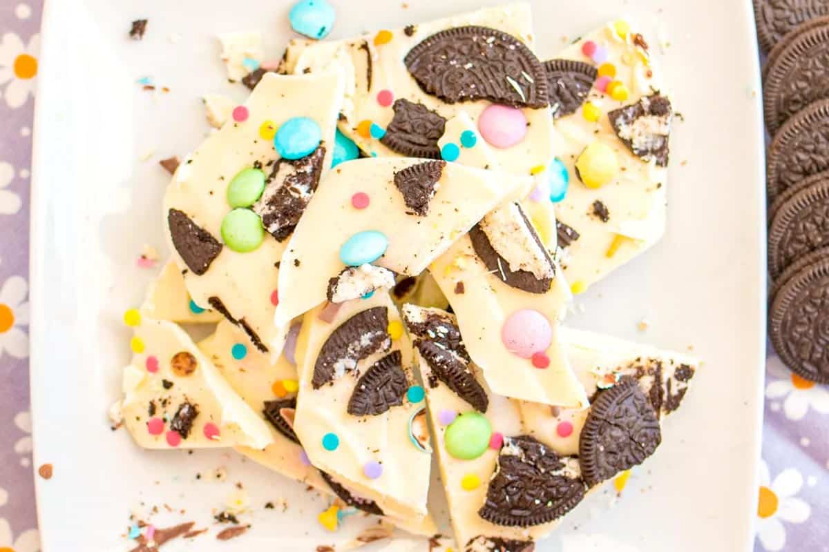 White chocolate bark topped with chocolate cookie pieces, colorful candy-coated chocolates, and sprinkles, arranged on a white plate.