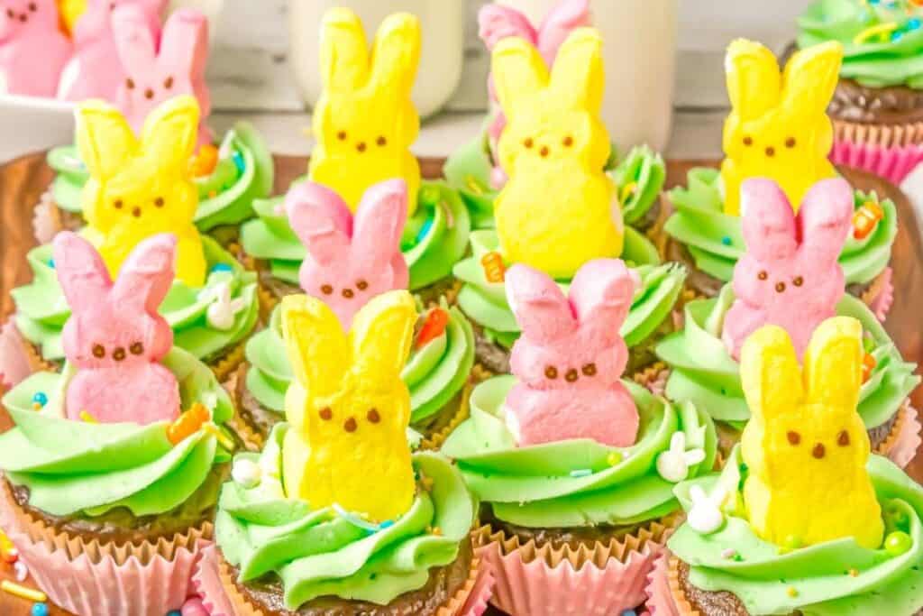 Cupcakes with green frosting are topped with pink and yellow marshmallow bunny candies and colorful sprinkles, arranged on a tray.