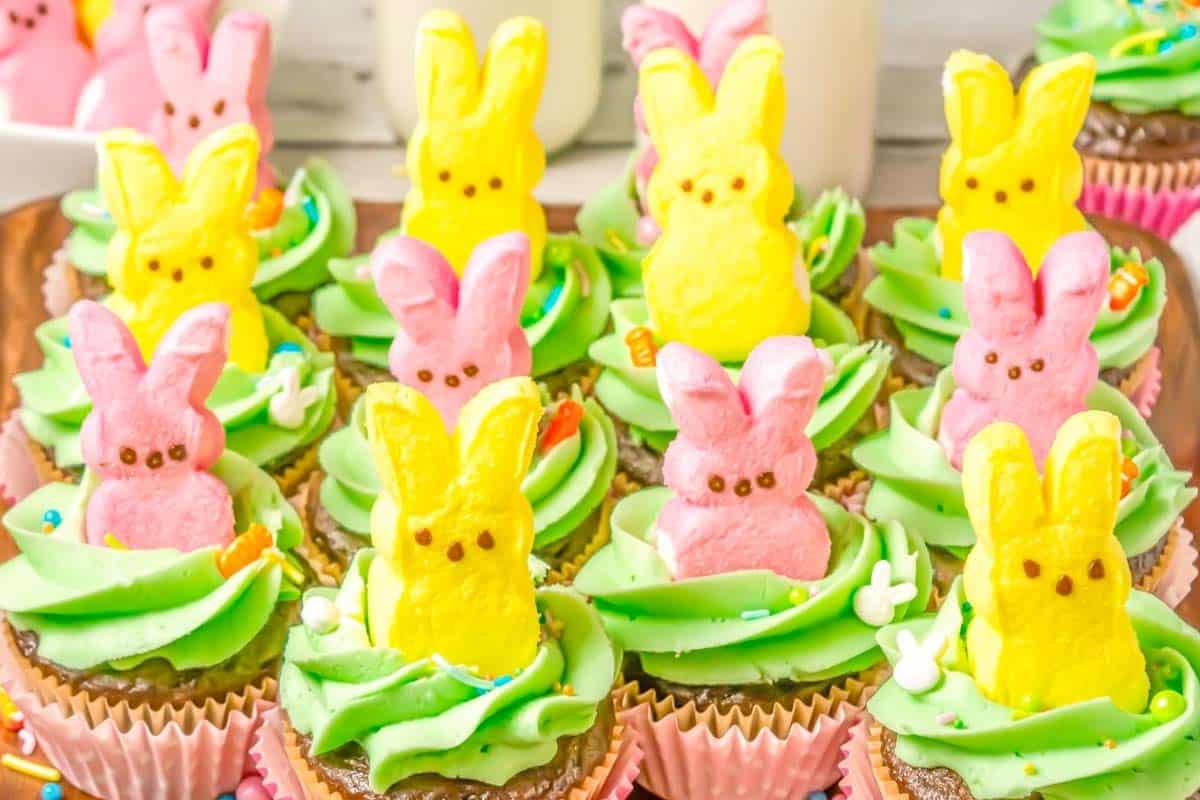Peeps Cupcakes with green frosting are topped with pink and yellow marshmallow bunny candies and colorful sprinkles, arranged on a tray.