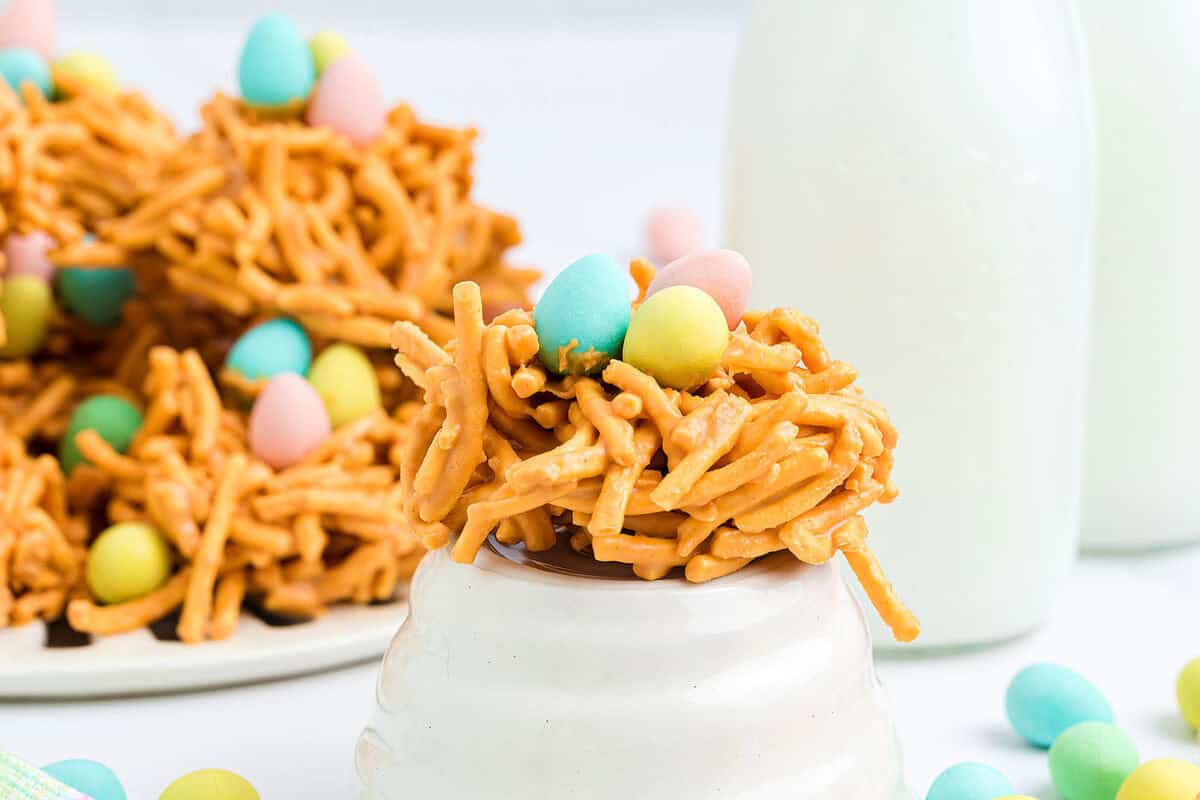 A no-bake treat made of chow mein noodles and butterscotch, shaped like a nest and topped with pastel candy eggs, is displayed in front of glass milk bottles.
