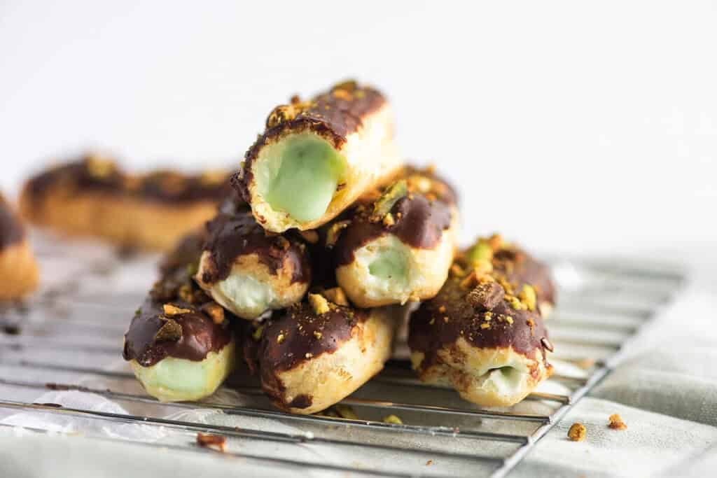A stack of chocolate-covered éclairs with a green pistachio filling sits on a wire cooling rack.