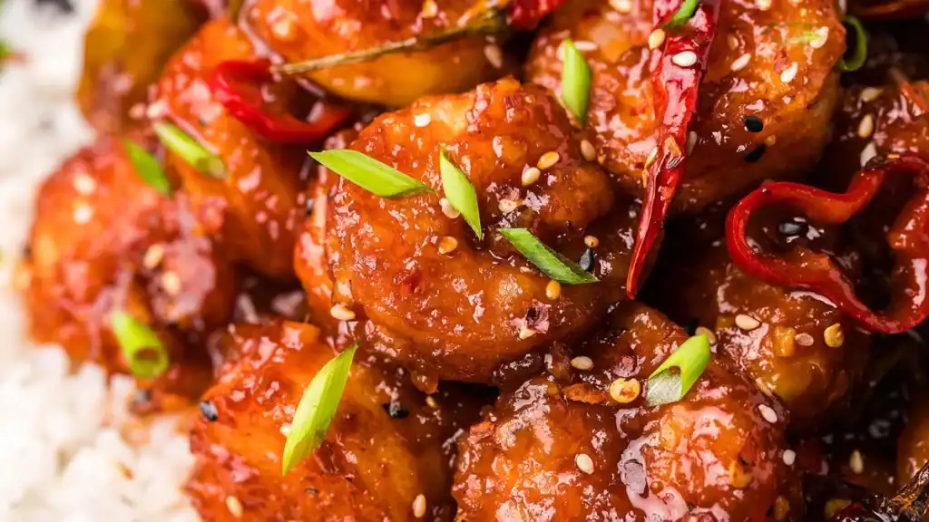 Close-up of spicy shrimp coated in a red sauce, garnished with sesame seeds, sliced green onions, and red chili peppers, served over a bed of white rice.