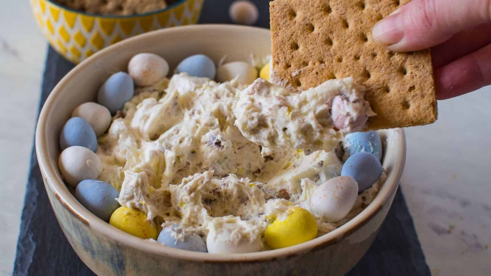 A hand dips a graham cracker into a bowl of creamy dip topped with candy-coated chocolate eggs, with more brown sugar in a bowl in the background.