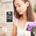A woman with long brown hair reads a menu at a table in a bright caf&eacute;, with rising restaurant prices on her mind. Purple flowers and blurred background details are visible.