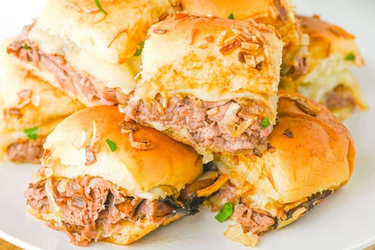 A plate of stacked French Dip Sliders on soft buns, topped with melted cheese and crispy onions, garnished with small green herbs.