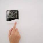A hand presses a button on a wall-mounted digital device displaying temperature, humidity, time, and date.