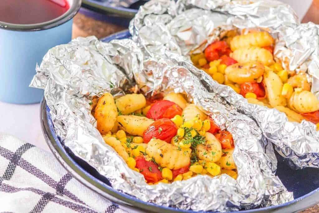 Foil packets filled with gnocchi, cherry tomatoes, corn, and herbs on a blue plate, next to a blue cup and a checkered cloth.