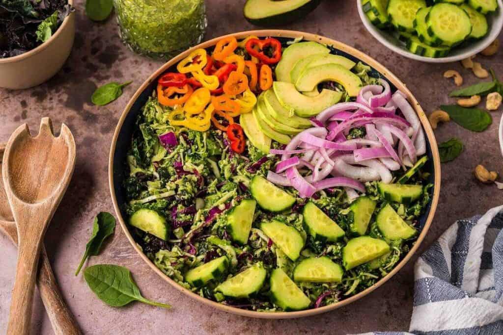 A salad bowl filled with sliced cucumbers, red onions, avocado, yellow and orange bell peppers, and mixed greens. Wooden salad tongs and other vegetables are nearby.