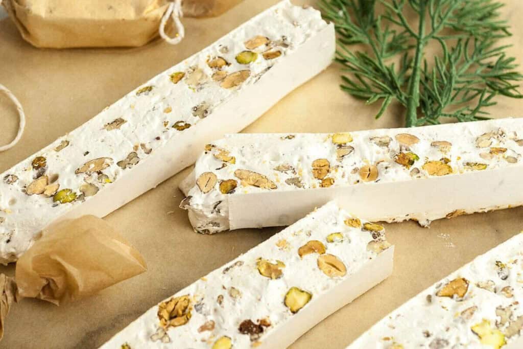Rectangular pieces of white nougat with visible nuts, placed on parchment paper next to a sprig of greenery and pieces of brown wrapped candy.