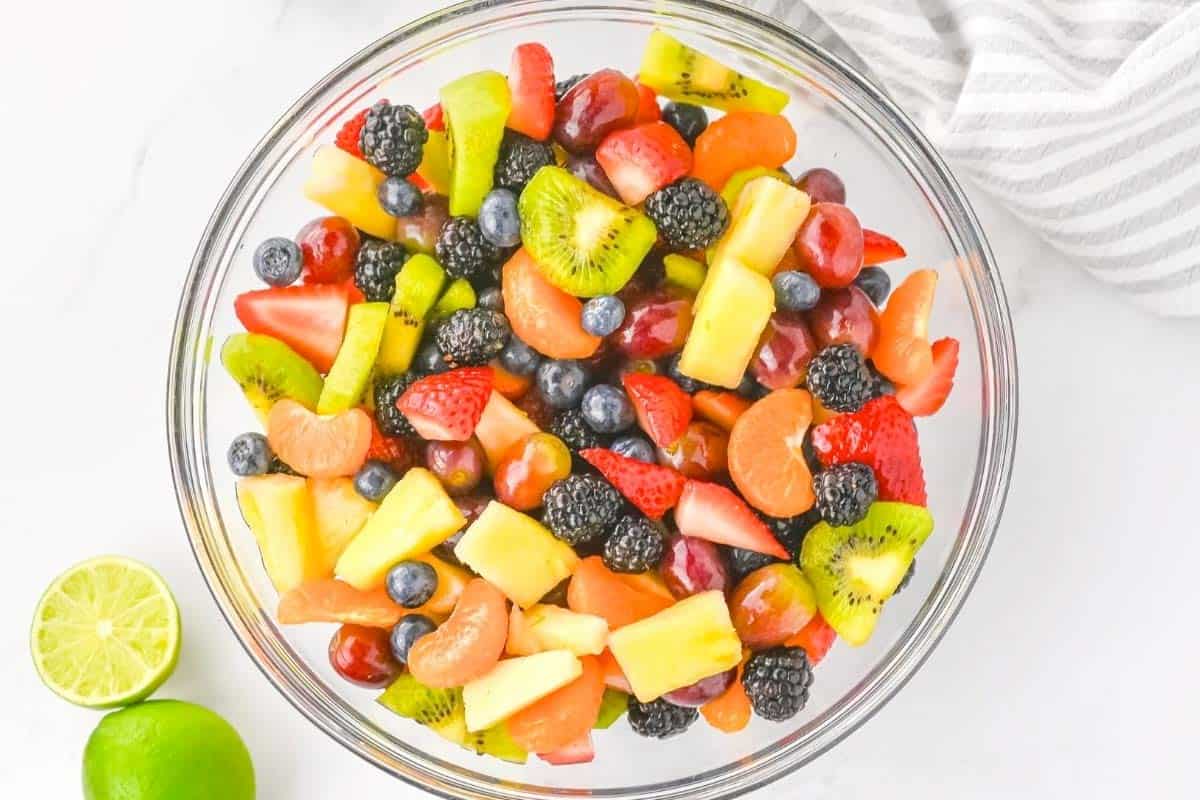 A glass bowl filled with assorted fresh fruit pieces including strawberries, kiwi, pineapple, grapes, blueberries, blackberries, and mandarin orange segments, with a halved lime nearby.