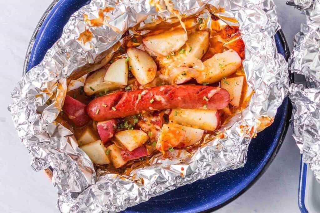 A foil packet containing a cooked sausage, diced potatoes, and vegetables, served in a blue dish.