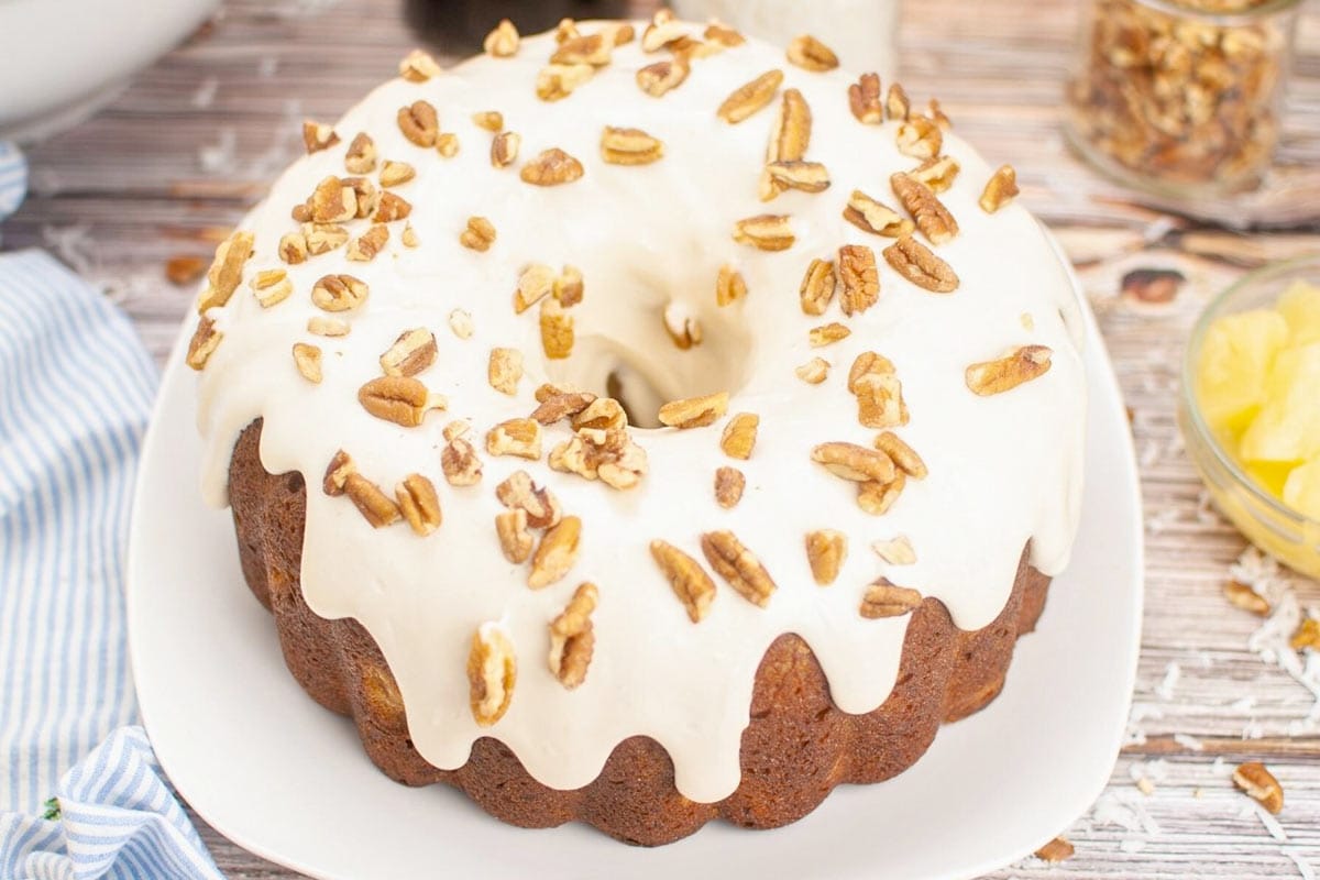 A bundt cake with white icing, topped with chopped pecans, sits on a white plate on a wooden surface.