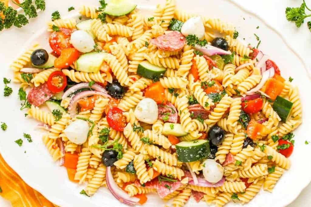 A platter of rotini pasta salad with cherry tomatoes, cucumbers, black olives, mozzarella balls, red onion, salami, bell peppers, and parsley.