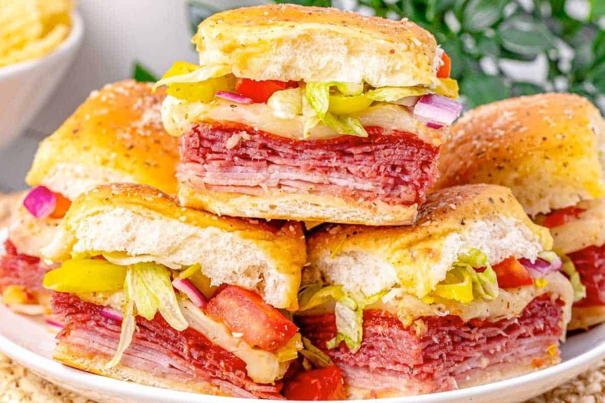 A plate of stacked Italian Sliders on seeded buns, filled with layers of sliced meats, cheese, lettuce, tomato, peppers, and red onion, with a green leafy plant and a bowl of potato chips in the background.