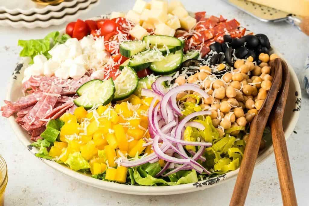 A large salad bowl filled with lettuce, sliced salami, cherry tomatoes, cubed cheese, cucumbers, chickpeas, olives, yellow peppers, red onions, and shredded cheese, with wooden salad tongs.