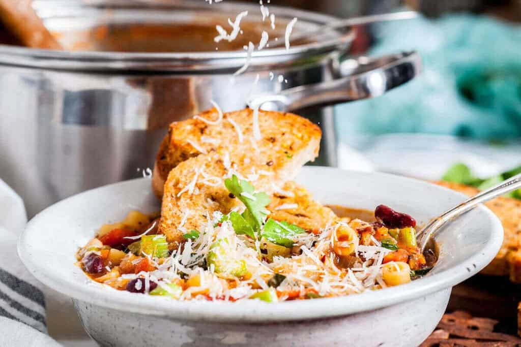 A bowl of vegetable soup with pasta and beans, topped with grated cheese and garnished with parsley, served with toasted bread slices. A pot is in the background.