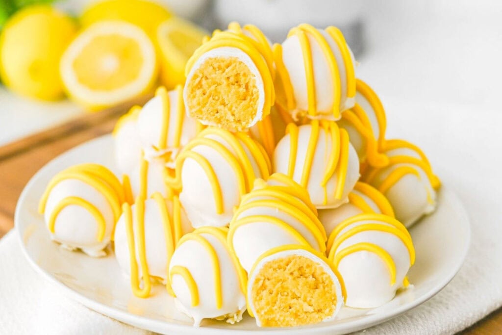 A plate of round lemon truffles coated in white chocolate with yellow drizzle, some with a bite showing the soft, yellow filling inside.
