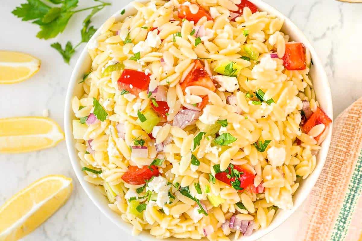 A bowl of orzo pasta salad with chopped tomatoes, red onions, celery, parsley, and feta cheese, placed on a marble surface next to lemon wedges and fresh parsley.