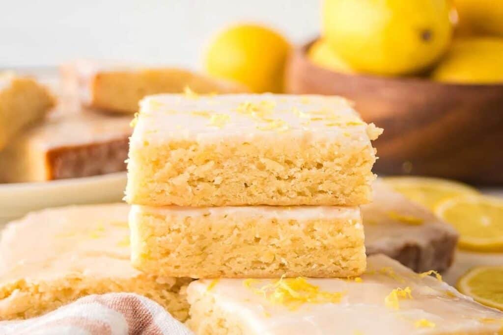 Two citrus desserts, lemon bars with icing, are stacked on top of each other, with more lemon bars and whole lemons in the background.