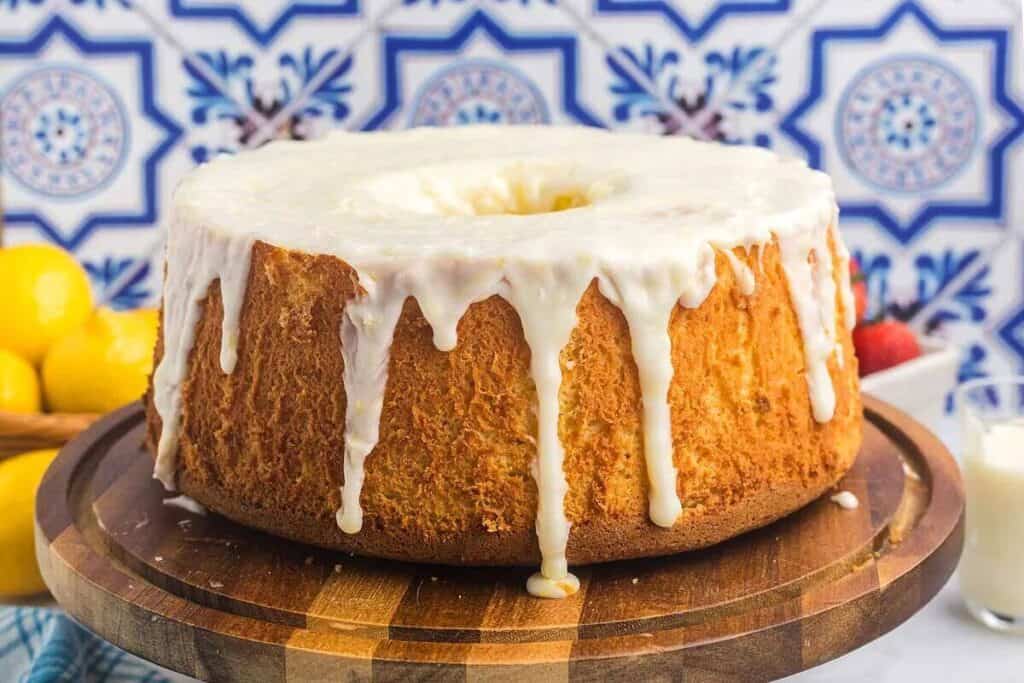 A glazed bundt-style cake with white icing drips, displayed on a wooden cake stand, highlights the charm of citrus desserts, with lemons, strawberries, and a glass of milk in the background.