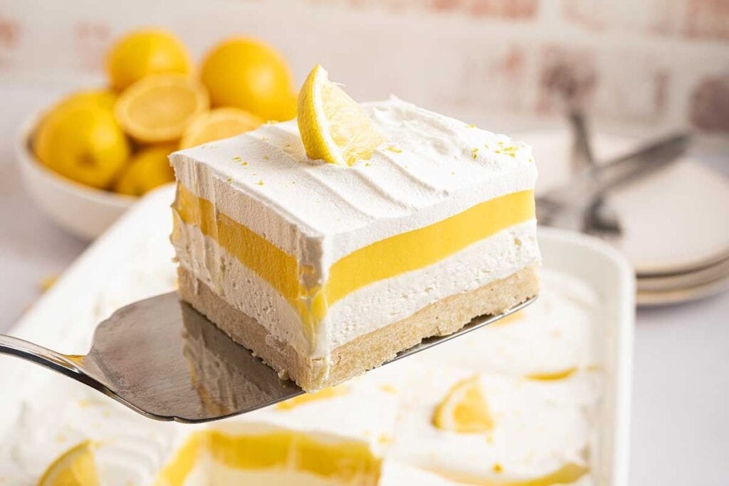 A spatula holds a square slice of layered lemon dessert with whipped topping and a lemon wedge, above a dish of similar pieces; lemons and plates are in the background.