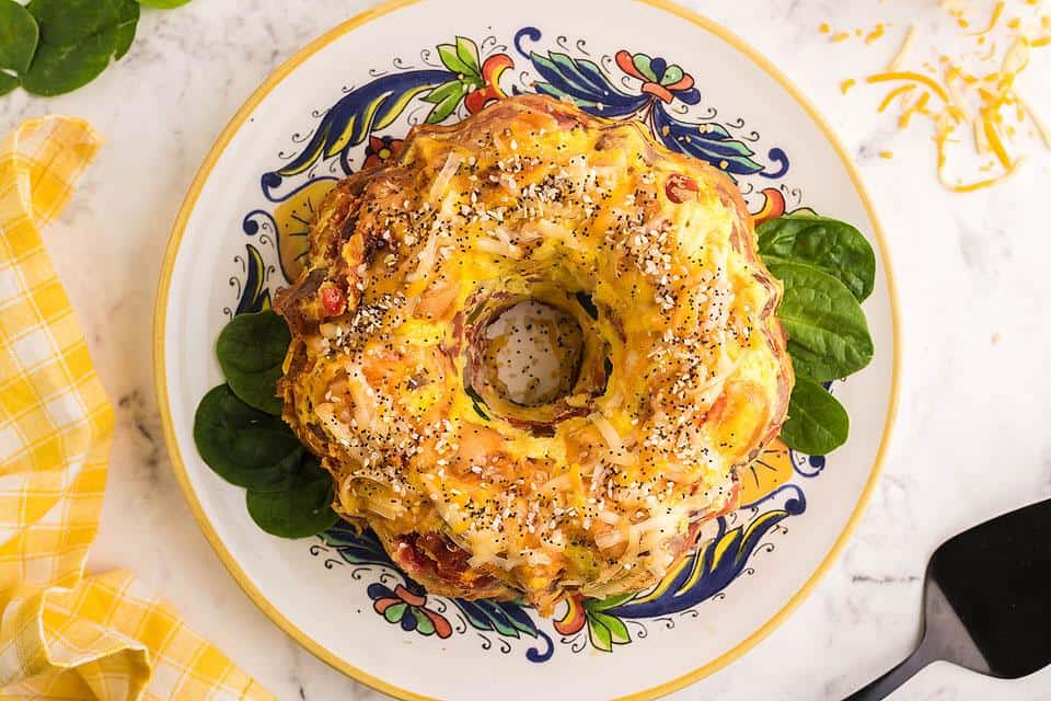 A savory bundt-shaped bread topped with shredded cheese and seeds sits on a decorative plate with spinach leaves underneath.