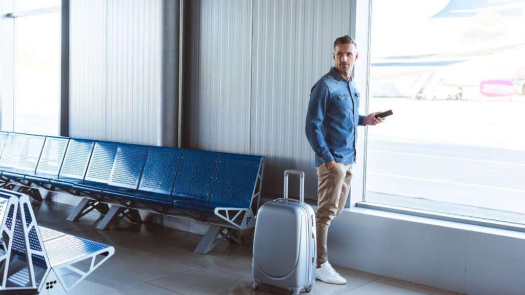 A man stands by a window in an airport terminal holding a phone, his suitcase beside him and empty blue seats nearby&mdash;perhaps checking his travel insurance before boarding.
