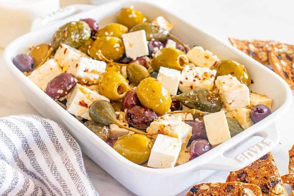 A white baking dish filled with mixed olives and cubes of feta cheese, garnished with herbs and spices, sits next to a striped napkin and crackers.