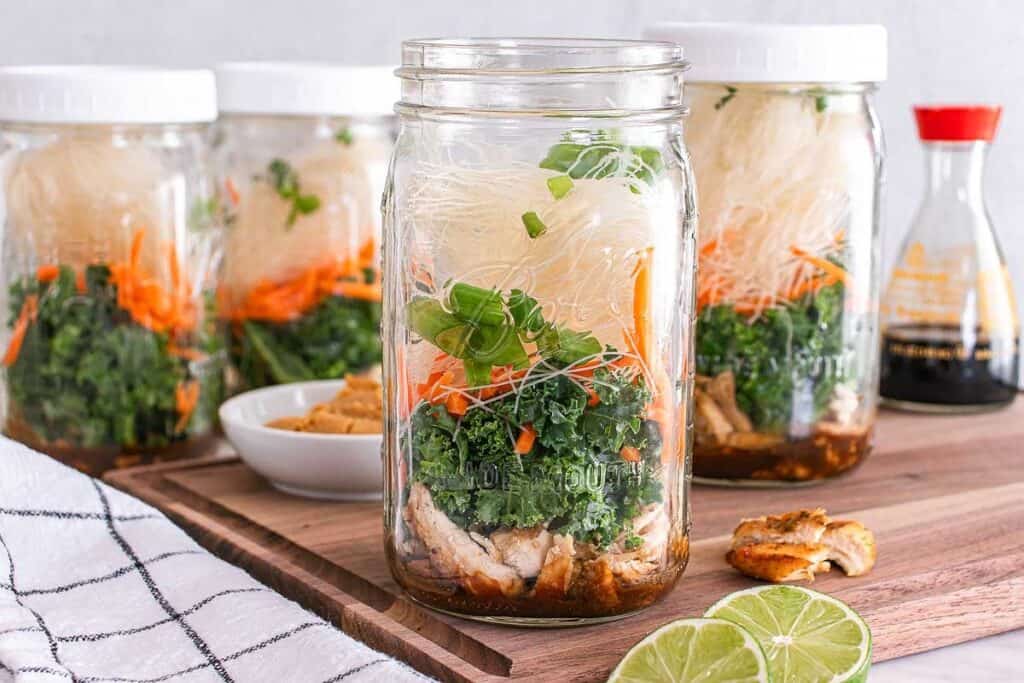 Four mason jars filled with layered ingredients for a noodle salad, including noodles, kale, carrots, and chicken, are placed on a wooden board near lime slices and a bottle of soy sauce.
