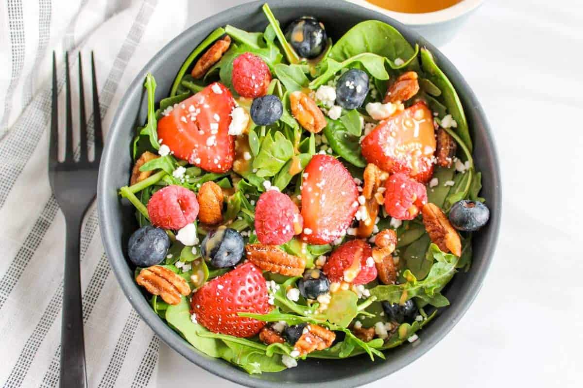 A bowl of spinach salad topped with strawberries, blueberries, raspberries, pecans, and crumbled cheese sits next to a fork and a cup of dressing.