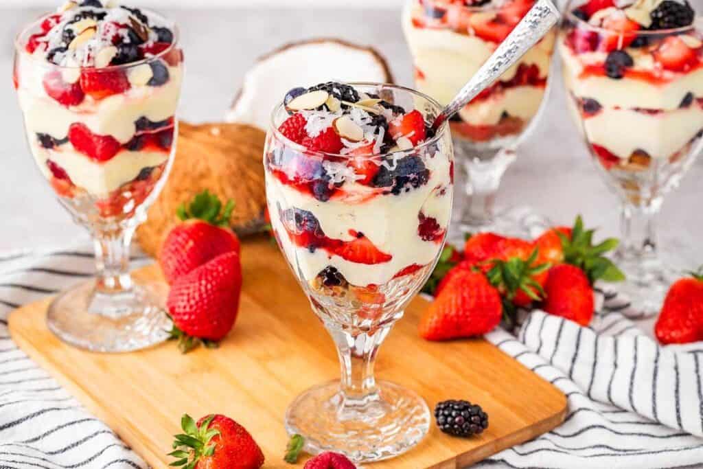 Four glass dessert cups filled with layers of custard, strawberries, blackberries, and blueberries are arranged on a wooden board with fresh berries scattered around.